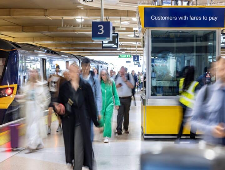 A bustling railway station platform with passengers in motion, featuring a blue and yellow train on the left and a yellow 'Customers with fares to pay' staff booth on the right.