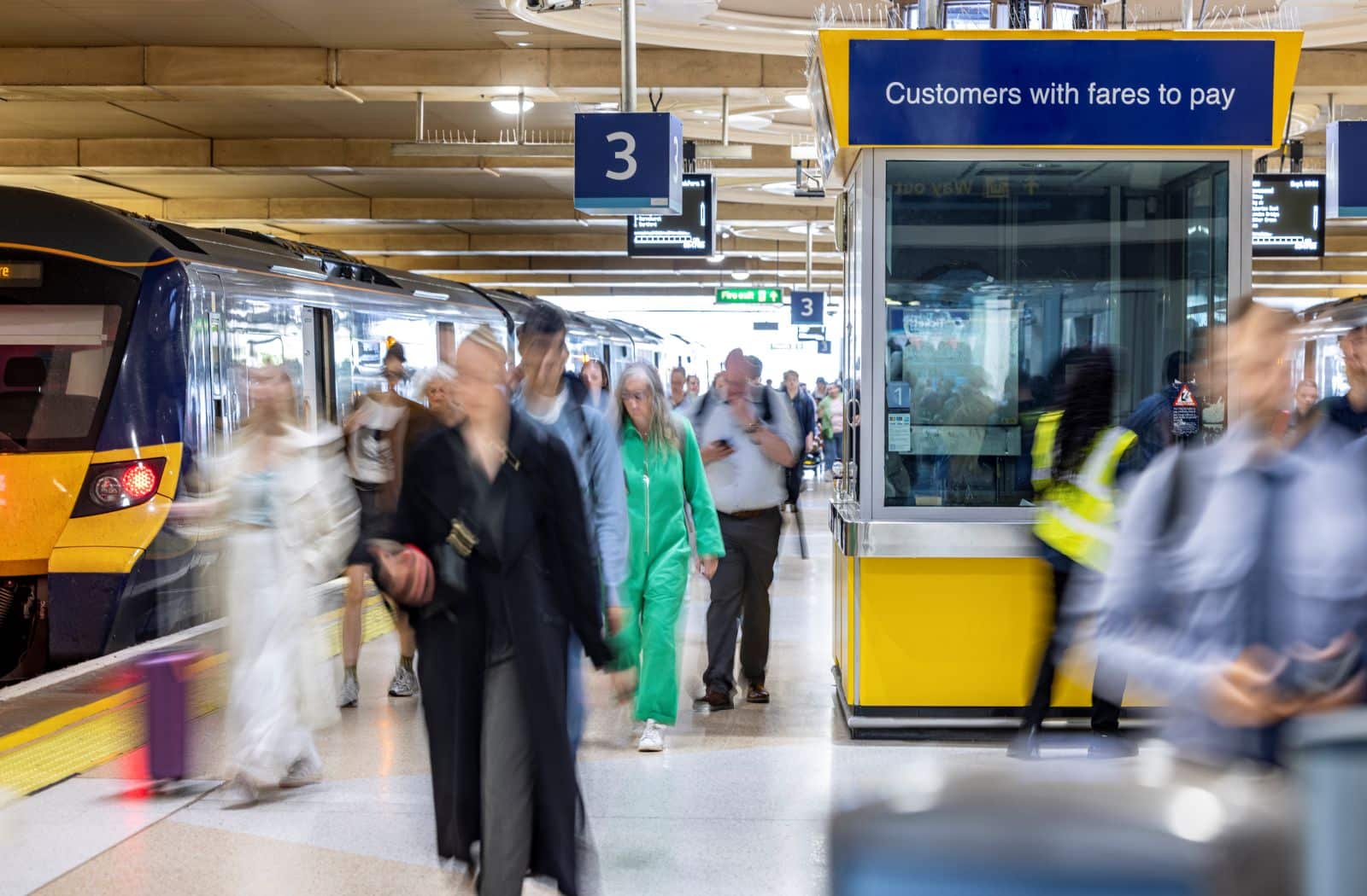 A bustling railway station platform with passengers in motion, featuring a blue and yellow train on the left and a yellow 'Customers with fares to pay' staff booth on the right.